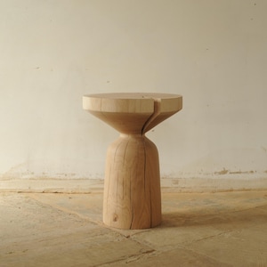 Könnte beinhalten: Ein Beistelltisch aus Naturholz mit einer einzigartigen Sanduhrform. Der Tisch hat eine runde Oberseite und einen ausgestellten, umgekehrt kegelförmigen Sockel. Die Holzmaserung ist sichtbar, und der Tisch ist etwa 46 cm hoch.