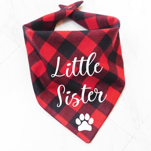 May include: Red and black plaid dog bandana with white text that says "Little Sister" and a white paw print.
