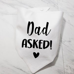 May include: White fabric bib with black text that reads "Dad Asked!" and a small black heart.