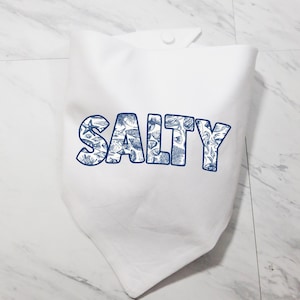 May include: White bandana with the word "SALTY" in blue, stylized text. The letters are filled with a detailed blue and white nautical pattern, including images of shells, starfish, and other sea life. The bandana is folded.