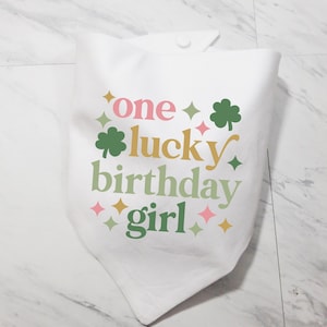 May include: White bandana with the text "one lucky birthday girl" in pink, green, yellow, and orange. The design includes shamrocks and star shapes. The bandana is folded and has a button closure.