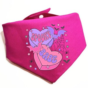 May include: Pink fabric bandana with a graphic of two candy hearts. The hearts are purple and pink with the text "Spooky Babe" in a retro font.