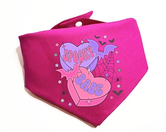 May include: Pink fabric bandana with a graphic of two candy hearts. The hearts are purple and pink with the text "Spooky Babe" in a retro font.