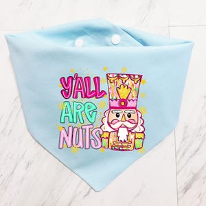 May include: Light blue pet bandana with a colorful graphic of a nutcracker and the text "Y'all Are Nuts". The bandana is made of soft fabric and has two white buttons.