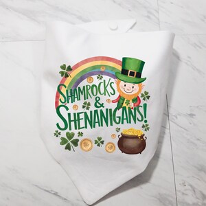 May include: White fabric with a green shamrock and rainbow design. The text "Shamrocks & Shenanigans!" is printed on the fabric. A cartoon leprechaun is holding a pot of gold.
