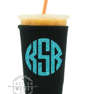 May include: A black insulated drink sleeve with a teal glitter monogram and a clear plastic cup filled with a beverage and an orange straw. The text "EAST ON TWENTY DESIGNS" is printed on the sleeve.