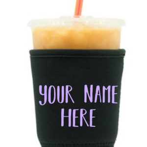 May include: A black fabric cup sleeve featuring the purple text "YOUR NAME HERE" printed on the front. The sleeve is wrapped around a clear plastic cup filled with a light brown beverage and includes a thin orange and white striped straw.