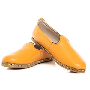 May include: A pair of yellow leather slip-on shoes with a brown stitched trim. The shoes have a slightly pointed toe and a flat sole.