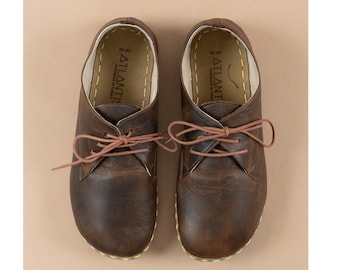 Op de afbeelding: Een paar bruine leren veterschoenen met een witte gestikte rand rond de zool. De schoenen hebben een bruine leren binnenzool met de tekst "M 40 ATLANTIS HANDMADE IN TURKEY" erop gedrukt.