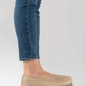 May include: Tan leather slip-on shoes with a stitched sole. The shoes are worn with blue jeans. The shoes have a minimalist design and a neutral color, suitable for various outfits.