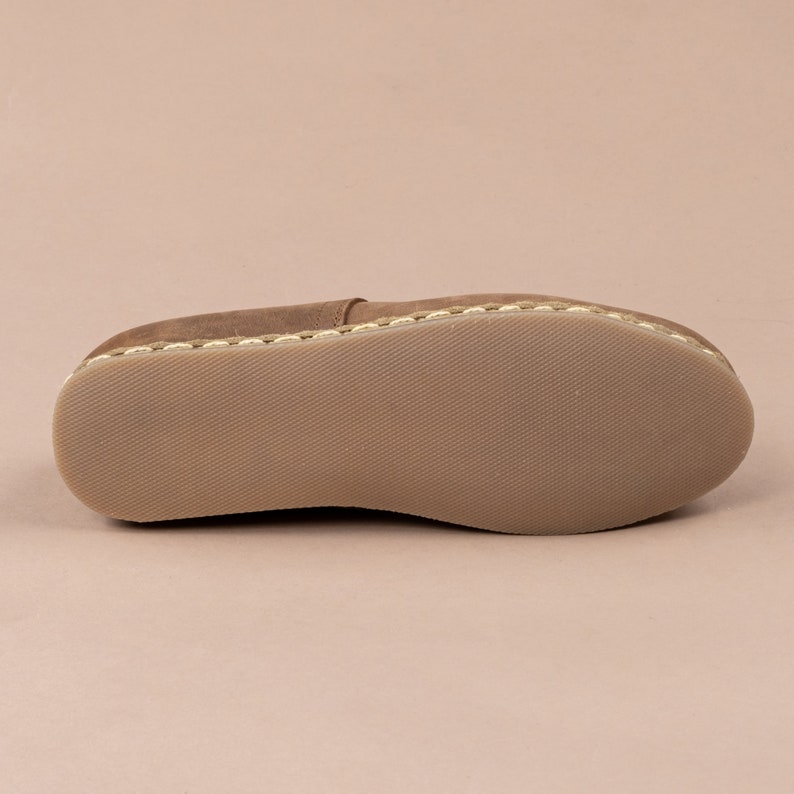 Puede incluir: La suela de un zapato mocas&iacute;n de cuero marr&oacute;n con un borde cosido blanco y una suela de goma texturizada.