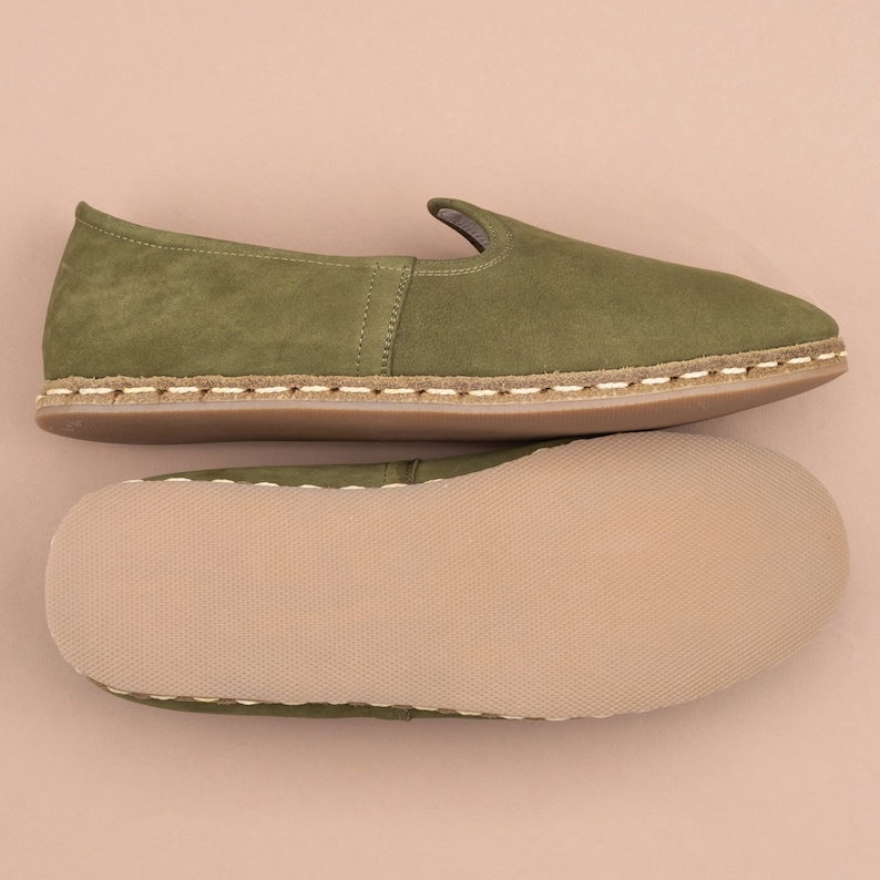 Pu&ograve; includere: Un paio di pantofole in camoscio verde con bordino bianco cucito e suola marrone. Le pantofole sono mostrate dall'alto e dal basso.