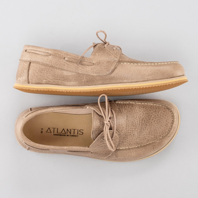 Pu&ograve; includere: Due paia di scarpe da barca beige con lacci, caratterizzate da un aspetto in pelle strutturata e cuciture bianche. Le scarpe hanno una suola marrone chiaro e il marchio "ATLANTIS" &egrave; visibile sulla soletta. Fatto a mano in Turchia.