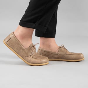 Pu&ograve; includere: Scarpe da barca color cuoio con tomaia in pelle strutturata e suola chiara. Le scarpe hanno un classico design allacciato e sono indossate con pantaloni neri. Le scarpe sono di colore neutro e adatte all'abbigliamento casual.