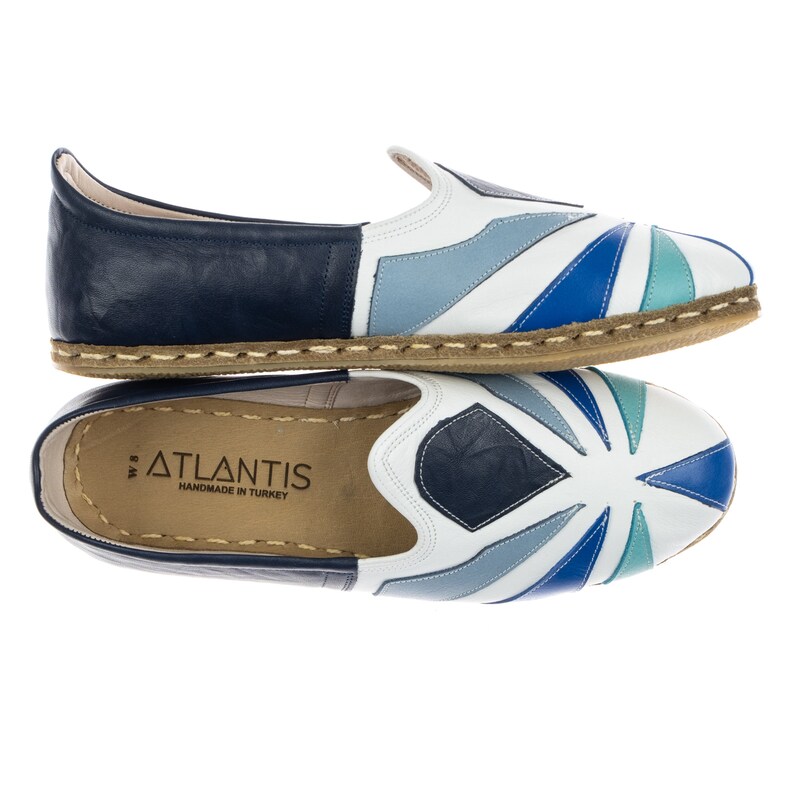 May include: A pair of white leather slip-on shoes with a blue, white, and teal geometric pattern. The shoes have a brown jute sole and the words "ATLANTIS HANDMADE IN TURKEY" are printed on the insole.