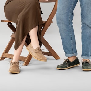 Pu&ograve; includere: Scarpe da barca in camoscio beige con lacci indossate da una persona seduta su una sedia di legno. Un'altra persona indossa scarpe da barca verdi con lacci. Entrambi indossano jeans. Stile classico e casual.