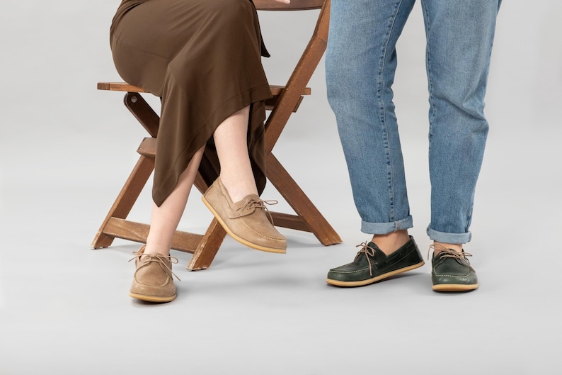 Pu&ograve; includere: Scarpe da barca in camoscio beige con lacci indossate da una persona seduta su una sedia di legno. Un'altra persona indossa scarpe da barca verdi con lacci. Entrambi indossano jeans. Stile classico e casual.