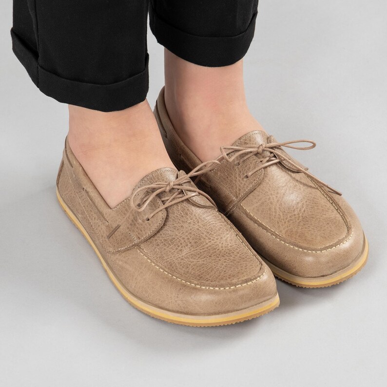 Pu&ograve; includere: Un paio di scarpe da barca beige con lacci. Le scarpe hanno un aspetto in pelle strutturata e una suola giallo chiaro. L'immagine mostra le scarpe da una prospettiva dall'alto, con i piedi di una persona che le indossa.