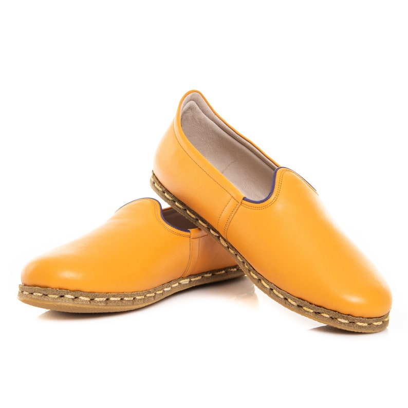 May include: A pair of yellow leather slip-on shoes with a brown stitched trim. The shoes have a slightly pointed toe and a flat sole.