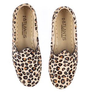 May include: A pair of leopard print slip-on shoes with a brown leather sole. The shoes have a label on the inside that reads "ATLANTIS HANDMADE IN TURKEY".