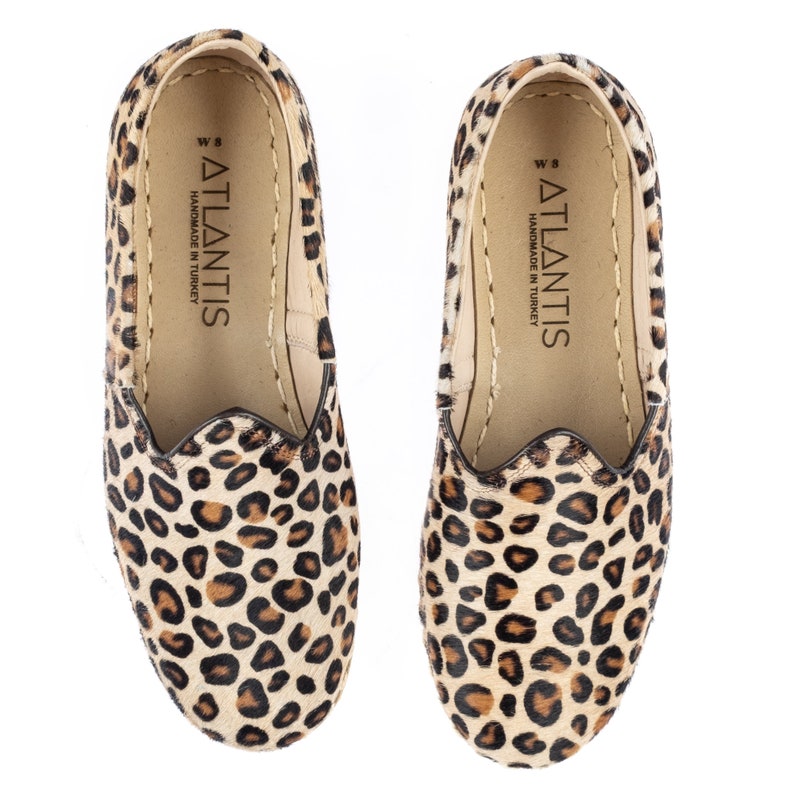 May include: A pair of leopard print slip-on shoes with a brown leather sole. The shoes have a label on the inside that reads "ATLANTIS HANDMADE IN TURKEY".
