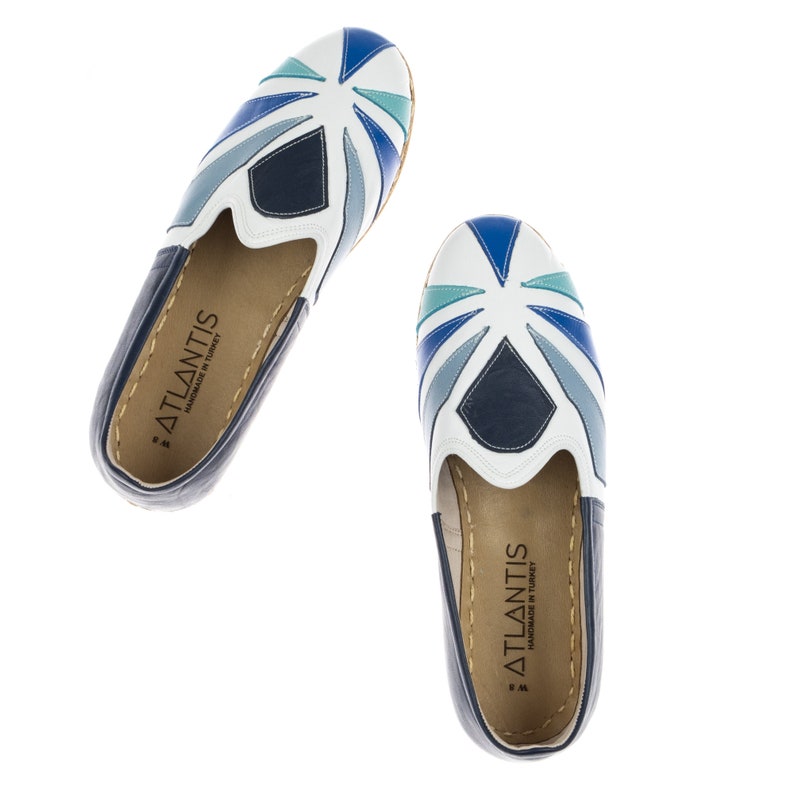 May include: A pair of white leather slip-on shoes with a blue and green geometric pattern. The shoes have a white sole and a blue leather lining. The shoes are handmade in Turkey and have the brand name "ATLANTIS" printed on the inside.