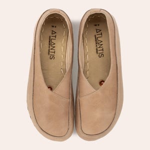 May include: A pair of tan leather slip-on shoes. The shoes have a rounded toe and a stitched sole. The brand name "ATLANTIS" is printed on the insole. The shoes are handmade in Turkey.