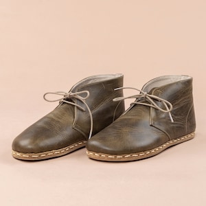 May include: A pair of olive green leather chukka boots with light-colored laces. The boots have a rounded toe and a stitched sole. The leather has a distressed appearance. The background is a solid, light peach color.