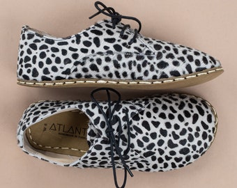 Può includere: Un paio di scarpe in pelle maculata bianca e nera con lacci neri. Le scarpe hanno una suola marrone chiaro e sono esposte su uno sfondo rosa chiaro. Taglia 8.