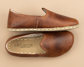 Könnte beinhalten: Ein Paar braune Leder-Slipper mit abgerundeter Zehenpartie und sichtbaren weißen Nähten entlang der Sohle. Die Innensohle zeigt den Text ATLANTIS HANDMADE IN TURKEY und die Größe W 8 in schwarzem Druck auf einem hellbeigen Hintergrund.