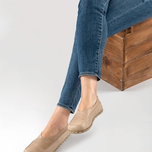 May include: Tan leather slip-on shoes with a rounded toe and a stitched sole. The shoes are worn with blue jeans. The shoes appear to be comfortable and casual.