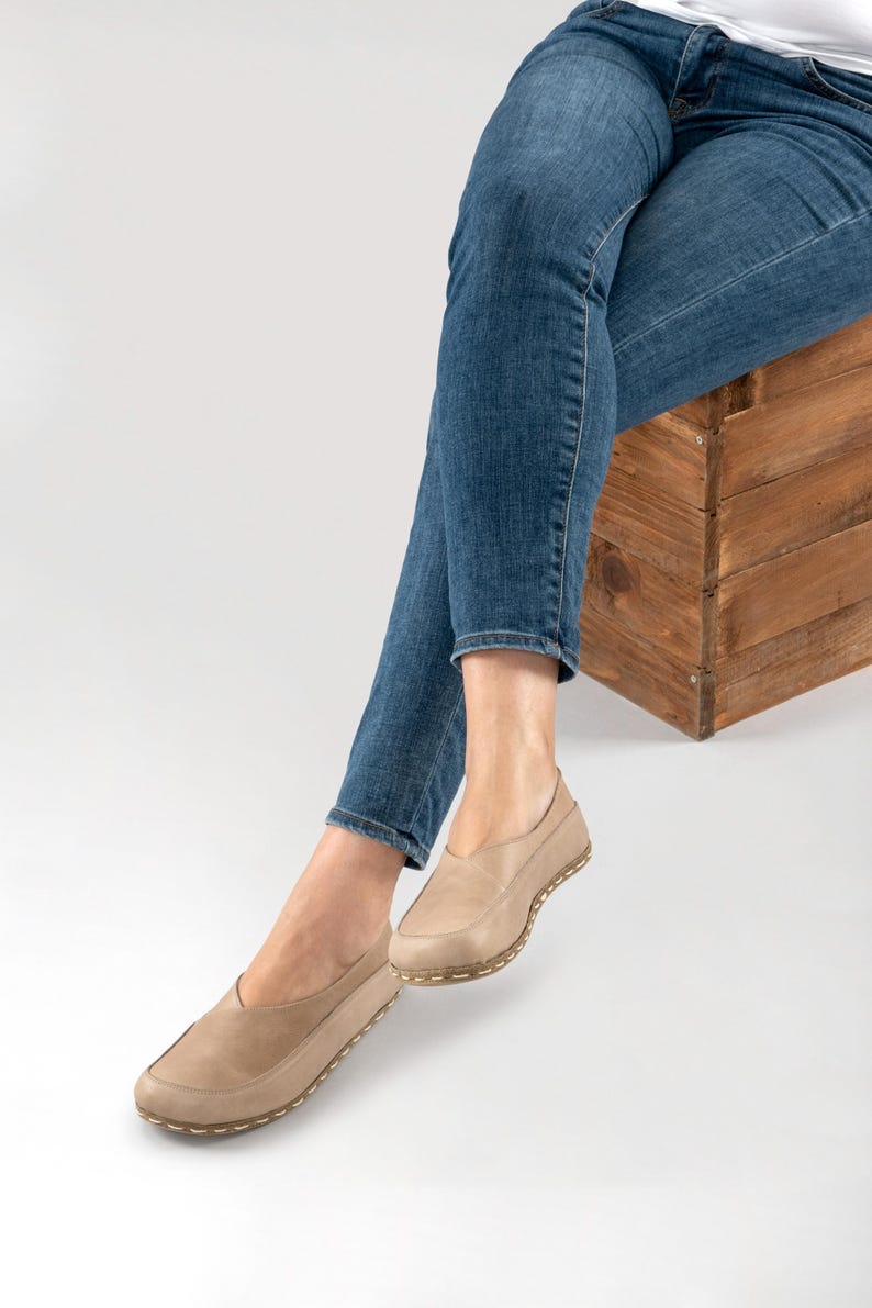 May include: Tan leather slip-on shoes with a rounded toe and a stitched sole. The shoes are worn with blue jeans. The shoes appear to be comfortable and casual.