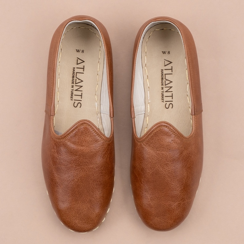 May include: A pair of brown leather slippers with a white stitched edge. The slippers have a white leather insole with the text "ATLANTIS HANDMADE IN TURKEY" and "W 8" printed on them.