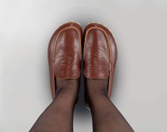 May include: A pair of brown leather loafers with a classic design. The shoes have a rounded toe and a stitched accent around the top. The shoes are worn with black sheer stockings. The background is a light gray.