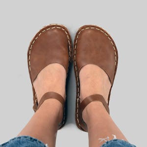Peut inclure: Une paire de chaussures en cuir marron avec un bout arrondi, une large bride sur le cou-de-pied et une fine bride de cheville avec boucle. Les chaussures présentent des coutures contrastantes blanches visibles le long de la semelle.