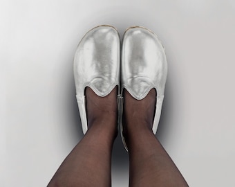 May include: A pair of silver slip-on shoes with a rounded toe and a decorative cutout design. The shoes are made of a shiny, metallic material and are shown from a top-down perspective.