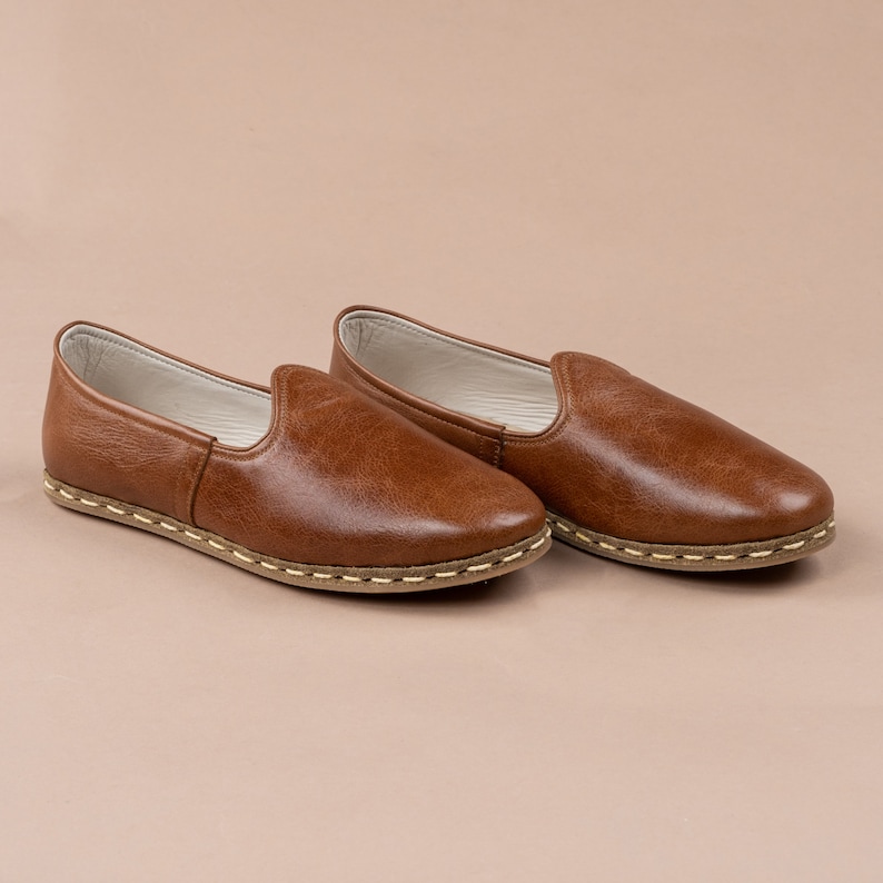 May include: A pair of brown leather slip-on shoes with a white stitch detail around the sole.