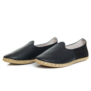 May include: A pair of black leather slip-on shoes with a light brown sole and stitching. The shoes have a simple design and are made of soft leather.