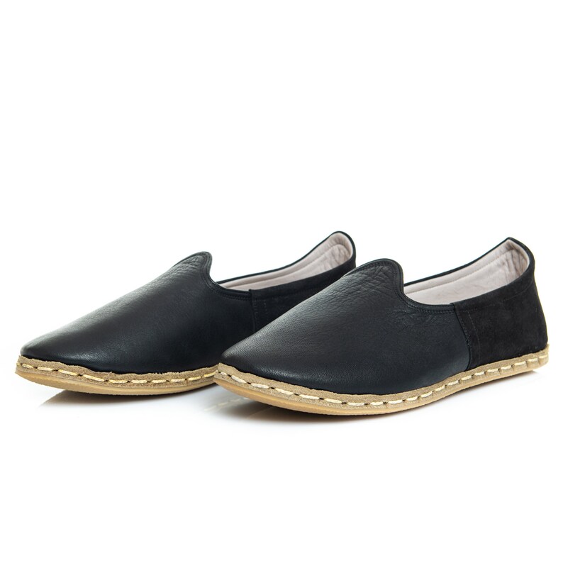 May include: A pair of black leather slip-on shoes with a light brown sole and stitching. The shoes have a simple design and are made of soft leather.