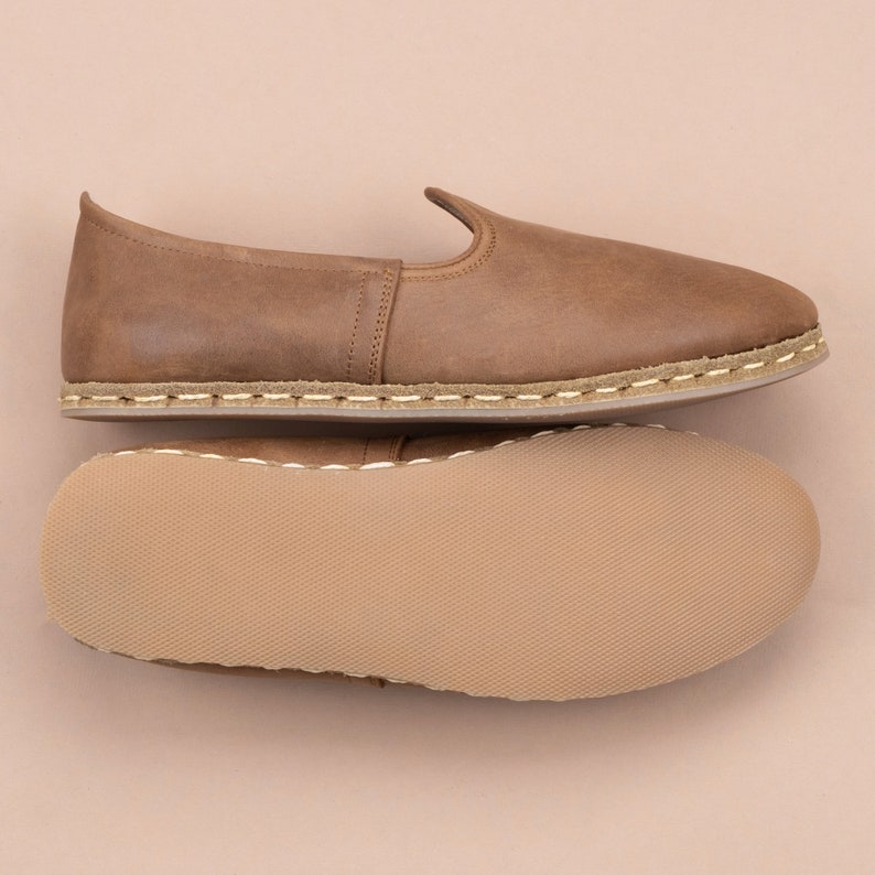 Puede incluir: Un par de zapatillas de cuero marr&oacute;n con un detalle de costura blanca alrededor de la suela. Las zapatillas se muestran desde arriba y abajo, revelando la plantilla.