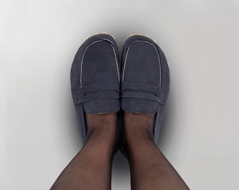 May include: A top-down view of two navy blue suede loafers. The shoes have a classic design with a strap detail across the top and white stitching. The soles are a light tan color.