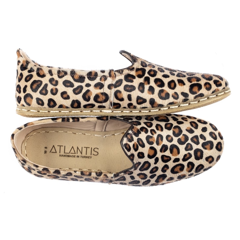 May include: A pair of tan leather slip-on shoes with a leopard print. The shoes have a white stitching detail around the sole and the words "ATLANTIS HANDMADE IN TURKEY" are printed on the insole.