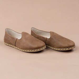Puede incluir: Un par de zapatos marrones de cuero sin cordones con un borde cosido y una suela marr&oacute;n claro.