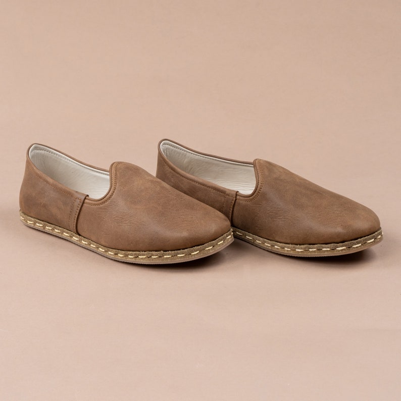Puede incluir: Un par de zapatos marrones de cuero sin cordones con un borde cosido y una suela marr&oacute;n claro.