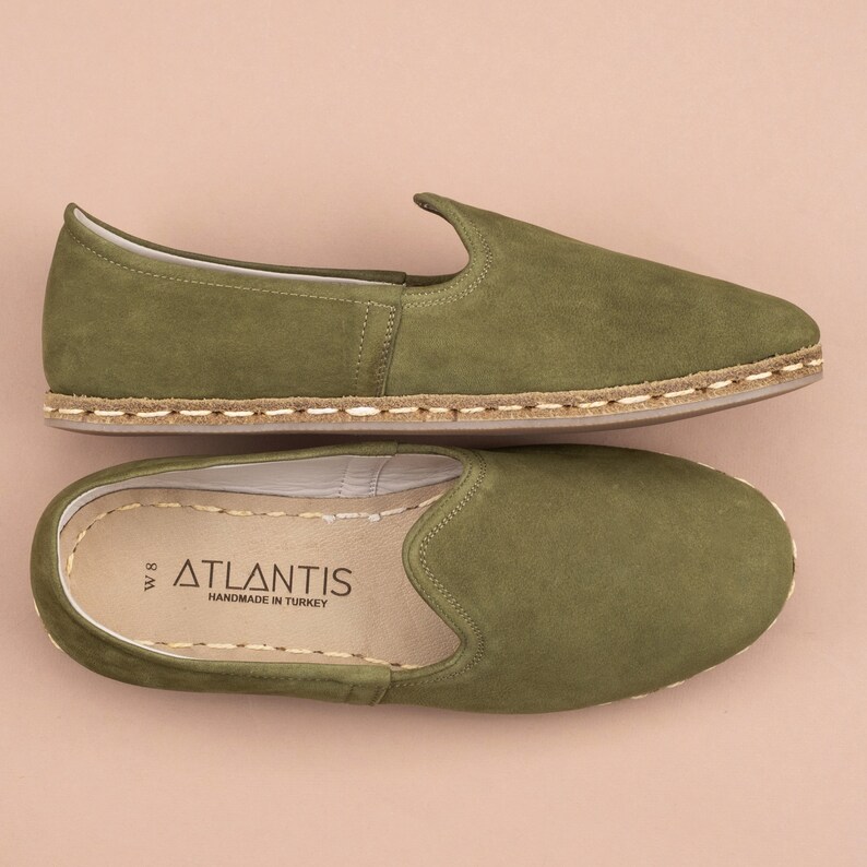 Pu&ograve; includere: Un paio di scarpe da uomo in pelle scamosciata verde con suola in juta. Le scarpe hanno un'etichetta con la scritta "ATLANTIS HANDMADE IN TURKEY".