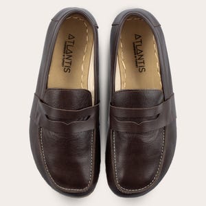 Può includere: Un paio di mocassini in pelle marrone con un dettaglio cucito attorno al bordo. I mocassini hanno una fodera in pelle e un dettaglio cucito bianco attorno alla parte superiore della scarpa. Le scarpe hanno il testo "ATLANTIS HANDMADE IN TURKEY" all'interno della scarpa.