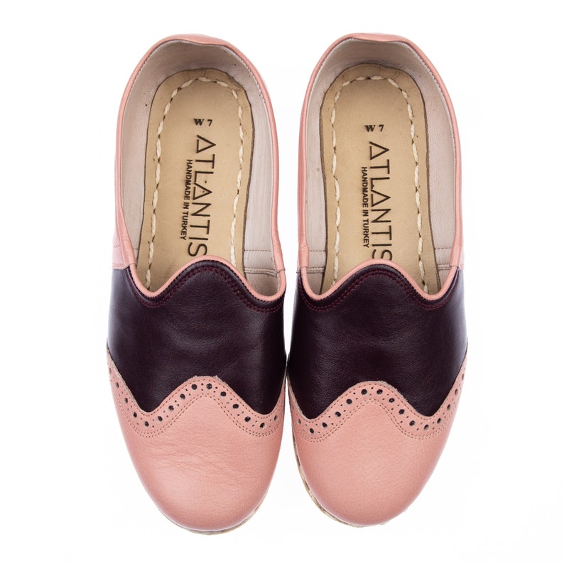 May include: A pair of pink and burgundy leather slip-on shoes with a wingtip design. The shoes are handmade in Turkey and have the brand name "ATLANTIS" printed on the inside.