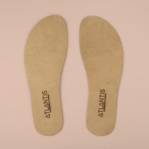 Könnte beinhalten: Zwei beige Leder-Einlegesohlen mit dem Text "ATLANTIS HANDMADE IN TURKEY" in Schwarz aufgedruckt. Die Einlegesohlen sind oval geformt und für den Einsatz in Schuhen konzipiert. Der Hintergrund ist hellrosa.