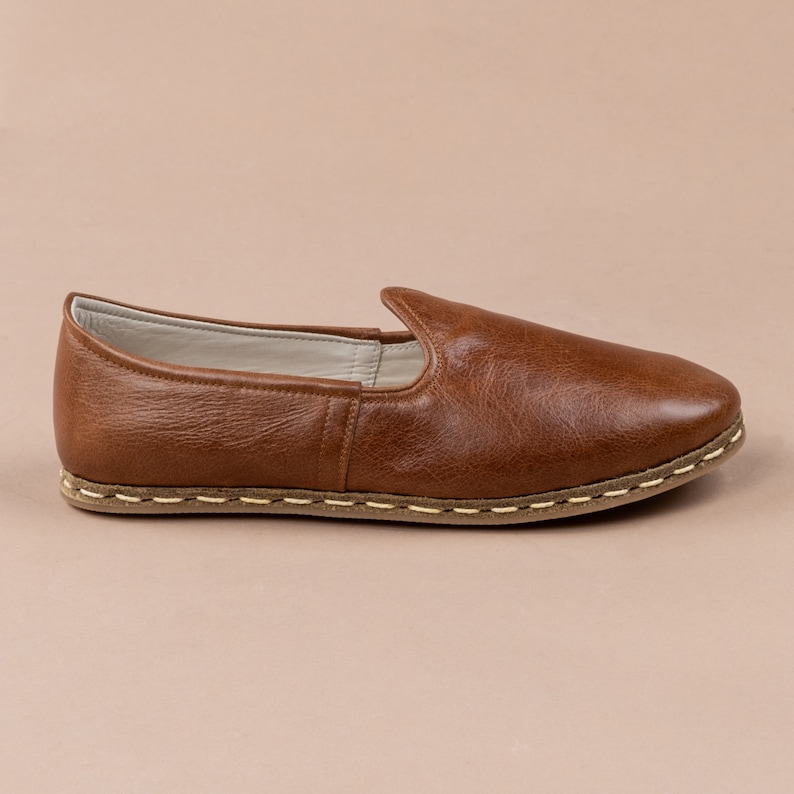 May include: A pair of brown leather slip-on shoes with a stitched edge and a light brown sole.