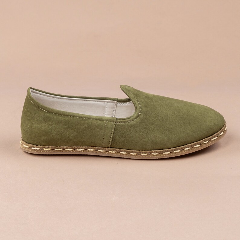Pu&ograve; includere: Un paio di scarpe da uomo in camoscio verde con suola marrone e bianca cucita.